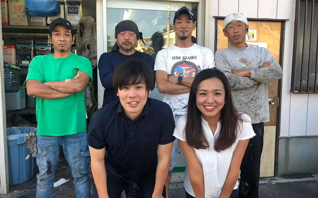 作業着姿の男性たちと私服の男女が屋外で笑顔を見せる集合写真