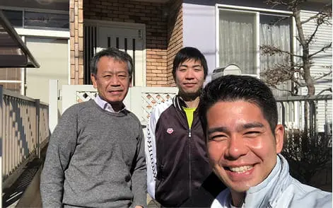 屋外で3人の男性が笑顔で並び、住宅の前で記念撮影している様子