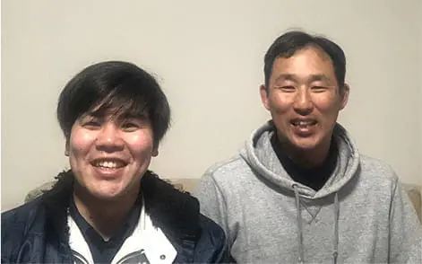 若い男性2人が室内でソファに座り、カメラに向かって笑顔を見せている