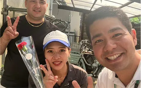 3人の男女が屋外で笑顔を見せ、1人の女性がカーネーションを手にしている