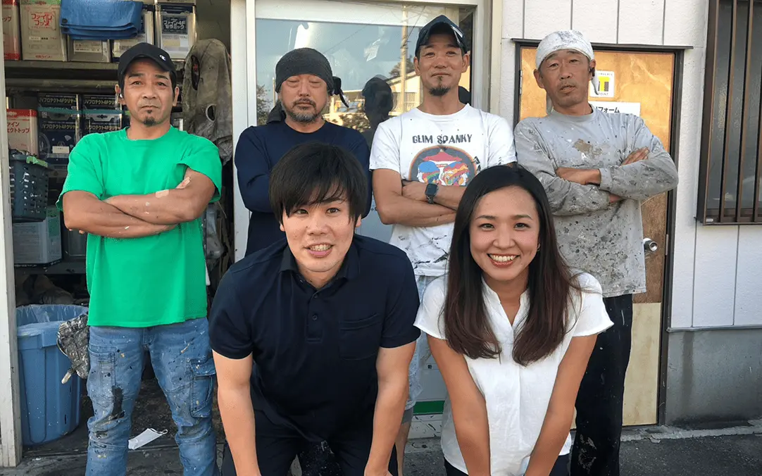 作業服姿の男性4人と私服の男女2人が店舗前で笑顔を見せる集合写真